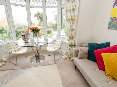 A sitting area with a round glass table and two white chairs near bay windows and a beige sofa with colorful cushions at Robin Hill in Duporth near St Austell