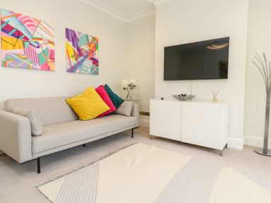 A living room with a beige sofa with colorful cushions a white cabinet with a flat screen TV above it two abstract paintings on the wall and a floor lamp at Robin Hill in Duporth near St Austell