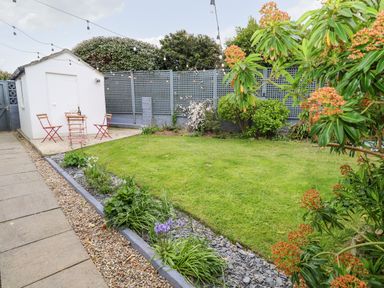 A garden with a lawn, flower beds, a small white shed, and a table with two chairs at Robin Hill in Duporth near St Austell