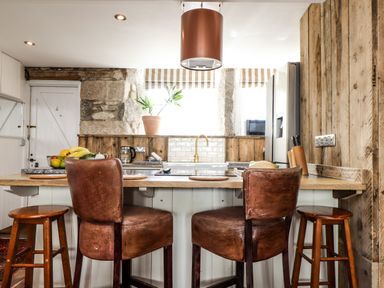 A kitchen with brown leather chairs around a wooden island and stone walls at Roncon's Villa in Goldsithney