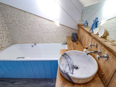 A bathroom with a white bathtub with blue paneling a round marble sink on a wooden countertop and a stone tile wall at Roncon's Villa in Goldsithney