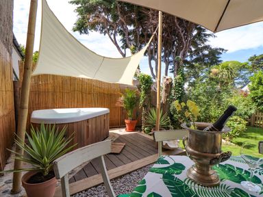 An outdoor patio area with a hot tub shaded by a white sail and a table with a bottle in an ice bucket at Roncon's Villa in Goldsithney