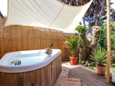 An outdoor jacuzzi with a champagne bucket and glasses under a sunshade with potted plants near a bamboo fence at Roncon's Villa in Goldsithney