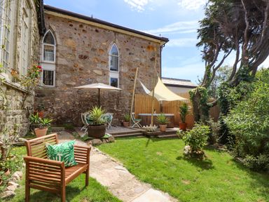 A garden with a wooden bench and a cushion a stone path potted plants an outdoor seating area with an umbrella a shade sail and a hot tub at Roncon's Villa in Goldsithney