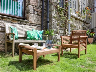 An outdoor seating area with wooden furniture including a bench with green leaf patterned cushions a chair and a low table with a pitcher and an open book against a stone wall at Roncon's Villa in Goldsithney
