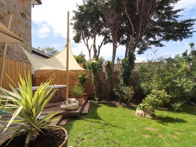A garden with grass plants trees a wooden deck with a sunshade and a hot tub at Roncon's Villa in Goldsithney