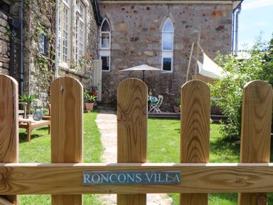 A wooden gate with a sign reading Roncons Villa opening to a garden with grass a stone path outdoor furniture and stone buildings in Goldsithney