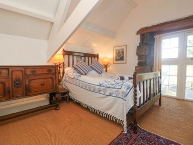 A bedroom with a wooden bed a patterned bedspread pillows a wooden dresser lamps a window and stone wall at Roncon's Villa in Goldsithney