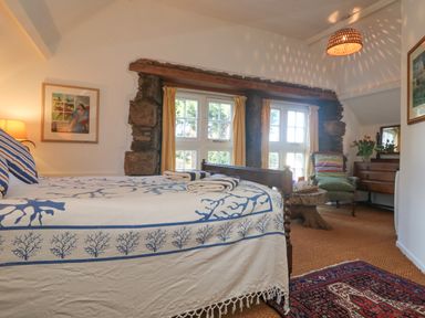 A bedroom with a bed covered by a patterned blanket a green armchair wooden dresser and a window with yellow curtains at Roncon's Villa in Goldsithney