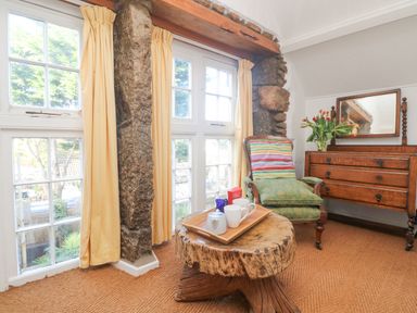 A room with large windows yellow curtains a wooden table with a tray a green armchair and a wooden dresser with flowers at Roncon's Villa in Goldsithney