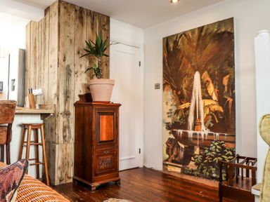 A corner of a room with wooden flooring a wooden cabinet with a potted plant a large painting of a fountain on the wall and wooden stools at Roncon's Villa in Goldsithney