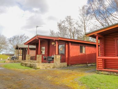 A group of red wooden cabins with stone patios and outdoor chairs in a natural setting at Ingram in Longframlington