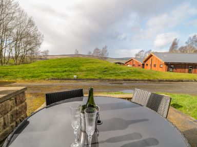 An outdoor patio table with two glasses and a bottle in a metal bucket with chairs and grassy hills and wooden buildings in the background at Ingram in Longframlington