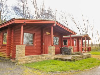 A red wooden cabin with stone pillars and an outdoor table with chairs at Ingram in Longframlington