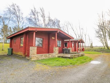 A red wooden cabin with stone foundation and outdoor table and chairs on a gravel driveway with trees in the background at Ingram in Longframlington