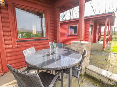 An outdoor patio area with a round glass table and four wicker chairs next to a red wooden cabin at Ingram in Longframlington