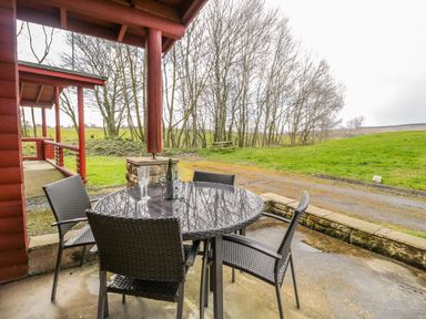 A patio with a round table and four chairs with a bottle and glass outside a red wooden cabin in a rural area at Ingram in Longframlington