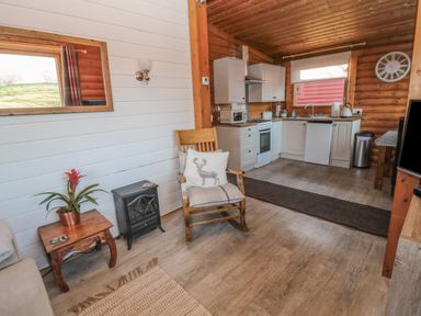 A wooden rocking chair with cushion in a living area next to a small table with a plant and a black heater near a kitchen with white cabinets at Ingram in Longframlington