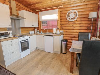 A kitchen with white cabinets a stove a microwave a kettle a sink a trash bin a dining table with black chairs and a wall clock at Ingram in Longframlington