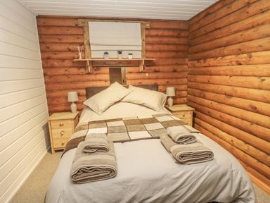 A bedroom with a bed made with beige pillows and a checkered blanket flanked by wooden nightstands with lamps and wooden log walls at Ingram in Longframlington