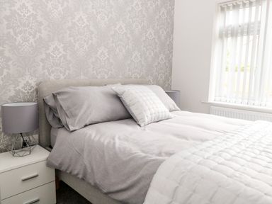 A bedroom with a bed grey bedding a pillow two bedside tables and lamps and a window with vertical blinds at Harbour Place in Wells-Next-The-Sea