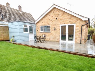 A backyard with green grass a tiled patio a small black metal table with two chairs and a brick house with glass doors at Harbour Place in Wells-Next-The-Sea