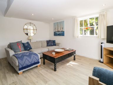A living room with a gray sofa with blue cushions a wooden coffee table a round wall mirror a painting of sailboats and a window with flowers at The Holt in Harcombe near Sidford