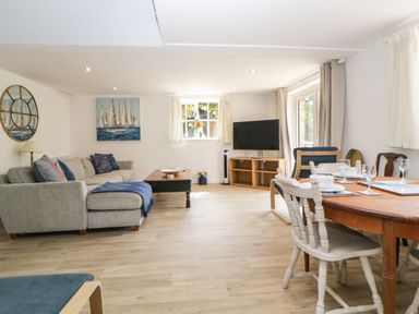 A living room with a gray sofa blue cushions a wooden coffee table a wooden dining table with white chairs a television on a stand and a window at The Holt in Harcombe near Sidford