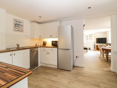 A kitchen with white cabinets wooden countertops a silver refrigerator and dishwasher leading to a dining area with a table chairs and a television at The Holt in Harcombe near Sidford