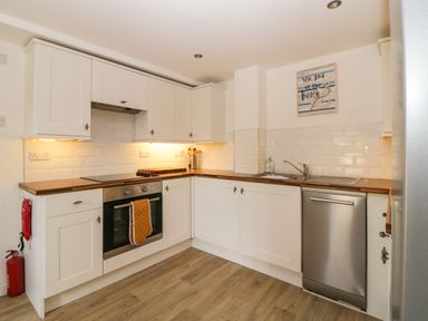 A kitchen with white cabinets wooden countertops an oven with a towel hanging on it a dishwasher a sink and a wall sign saying see you at the beach at The Holt in Harcombe near Sidford