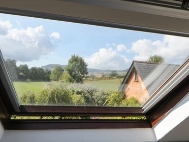 A view of green fields trees and a brick building roof through an open skylight window at The Holt in Harcombe near Sidford