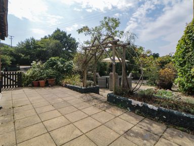 A tiled patio with wooden pergola and outdoor seating surrounded by plants and trees at The Holt in Harcombe near Sidford