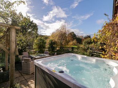An outdoor patio with a hot tub and a table with four wicker chairs surrounded by trees and plants at The Holt in Harcombe near Sidford