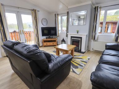 A living room with two black leather sofas a wooden coffee table a TV on a wooden stand a fireplace and windows with curtains at Lavender Lodge Applethwaite 4 Limefitt Park near Windermere