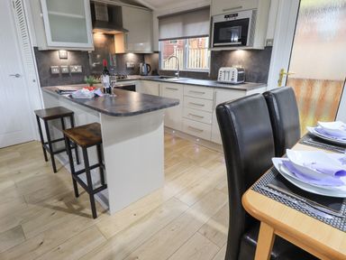 A kitchen with an island counter, bar stools, sink, built-in microwave, toaster, wine bottle and glasses near a dining table with two chairs at Lavender Lodge Applethwaite 4 Limefitt Park near Windermere
