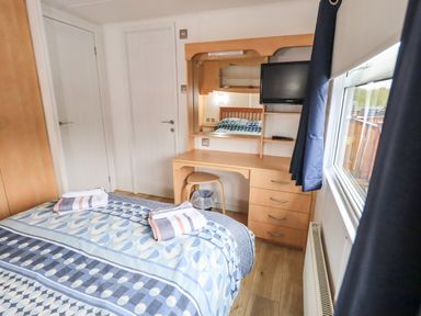 A bedroom with a bed, a dresser with a mirror, a stool, two closed doors, a window with curtains, a radiator, and a television at Lavender Lodge in Applethwaite 4 Limefitt Park near Windermere
