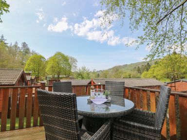 An outdoor wooden deck with a round glass table and four wicker chairs with a bottle and glasses on the table at Lavender Lodge Applethwaite 4 Limefitt Park near Windermere