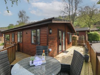 An outdoor wooden deck with a glass table and wicker chairs outside a wooden cabin at Lavender Lodge in Applethwaite 4 Limefitt Park near Windermere