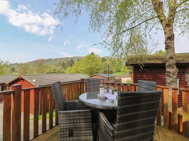 A wooden deck with a round glass table and four woven chairs with a bottle and glasses on the table at Lavender Lodge Applethwaite 4 Limefitt Park near Windermere