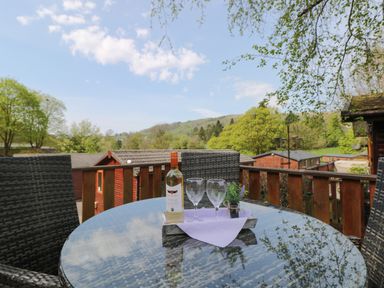 An outdoor patio table with a bottle of wine two wine glasses and a small plant with trees and rooftops in the background at Lavender Lodge Applethwaite 4 Limefitt Park near Windermere