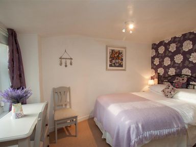 A bedroom with a bed covered by a purple blanket a white desk with a chair and a vase with flowers a wall decorated with floral wallpaper at Rustic Cottage in Bowness-On-Windermere