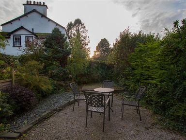 An outdoor seating area with a round table and four metal chairs surrounded by greenery and a white house in the background at Rustic Cottage in Bowness-On-Windermere