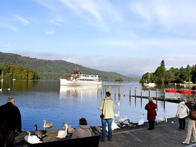 People standing and sitting by a lake with swans and a large boat on the water at Rustic Cottage in Bowness-On-Windermere