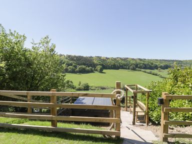 A wooden fence and gate with a rope handle leading to a tiled patio with a bench and green fields and trees in the background at Kaitkyella Lodge in Scarborough