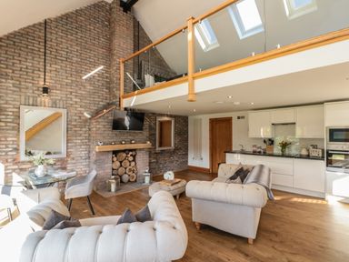A living room with beige sofas glass table kitchen area and brick wall with stacked logs at Kaitkyella Lodge in Scarborough