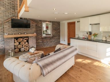 An open plan living room with a beige sofa a wood stacked fireplace a wall mounted tv kitchen cabinets and a wooden floor at Kaitkyella Lodge in Scarborough