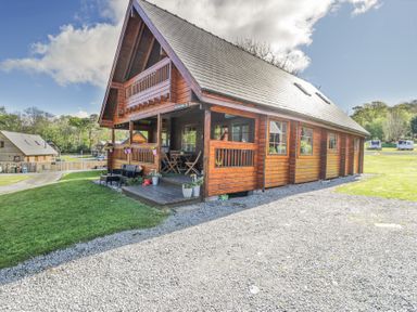 A wooden lodge with a covered porch and outdoor seating on a gravel and grass area at Sun View Lodge in Llanbedr