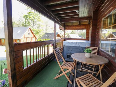 A covered wooden balcony with a round table wooden chairs a hot tub and wooden houses in the background at Sun View Lodge in Llanbedr