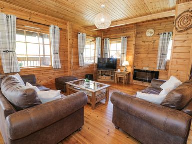 A living room with brown sofas, a glass coffee table, a television, and windows with curtains at Sun View Lodge in Llanbedr