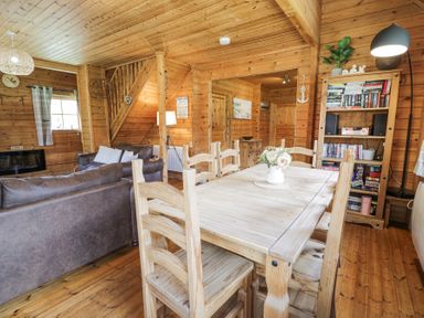 A wooden dining table with six chairs and a flower vase in a wooden walled living room with sofas and a bookshelf at Sun View Lodge in Llanbedr